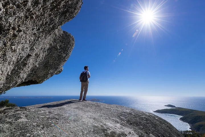 Persona con mochila de pie sobre una gran roca con vistas al océano bajo un sol brillante.