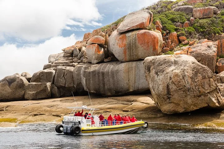 Barco anfibio amarillo con pasajeros explorando cerca de grandes rocas de granito cubiertas de líquenes naranjas.